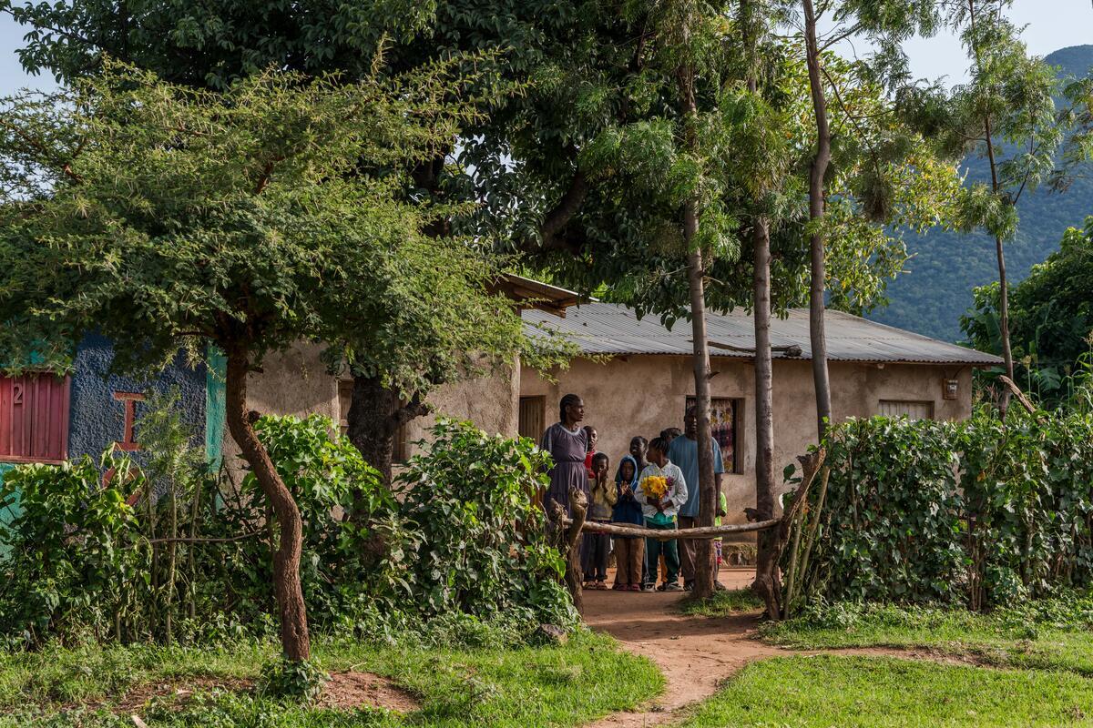 A group of people stand outside a modest rural home, surrounded by lush greenery, trees, and a simple fence on a sunny day