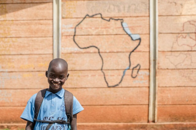 A smiling boy in a school uniform and backpack stands in front of a wall painted with an outline map of Africa.