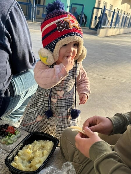 A young girl puts food in her mouth while watching an adult who is making bite-size pieces for her. There is a building behind her and another adult sitting next to her.