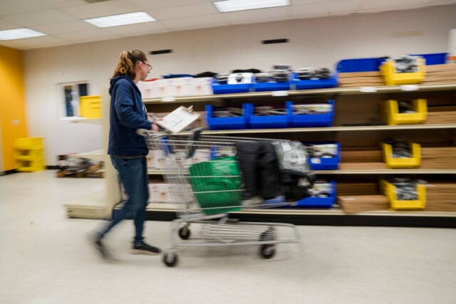 A woman pushes a shopping cart filled with items while holding a clipboard. She is looking at shelves of classroom materials.