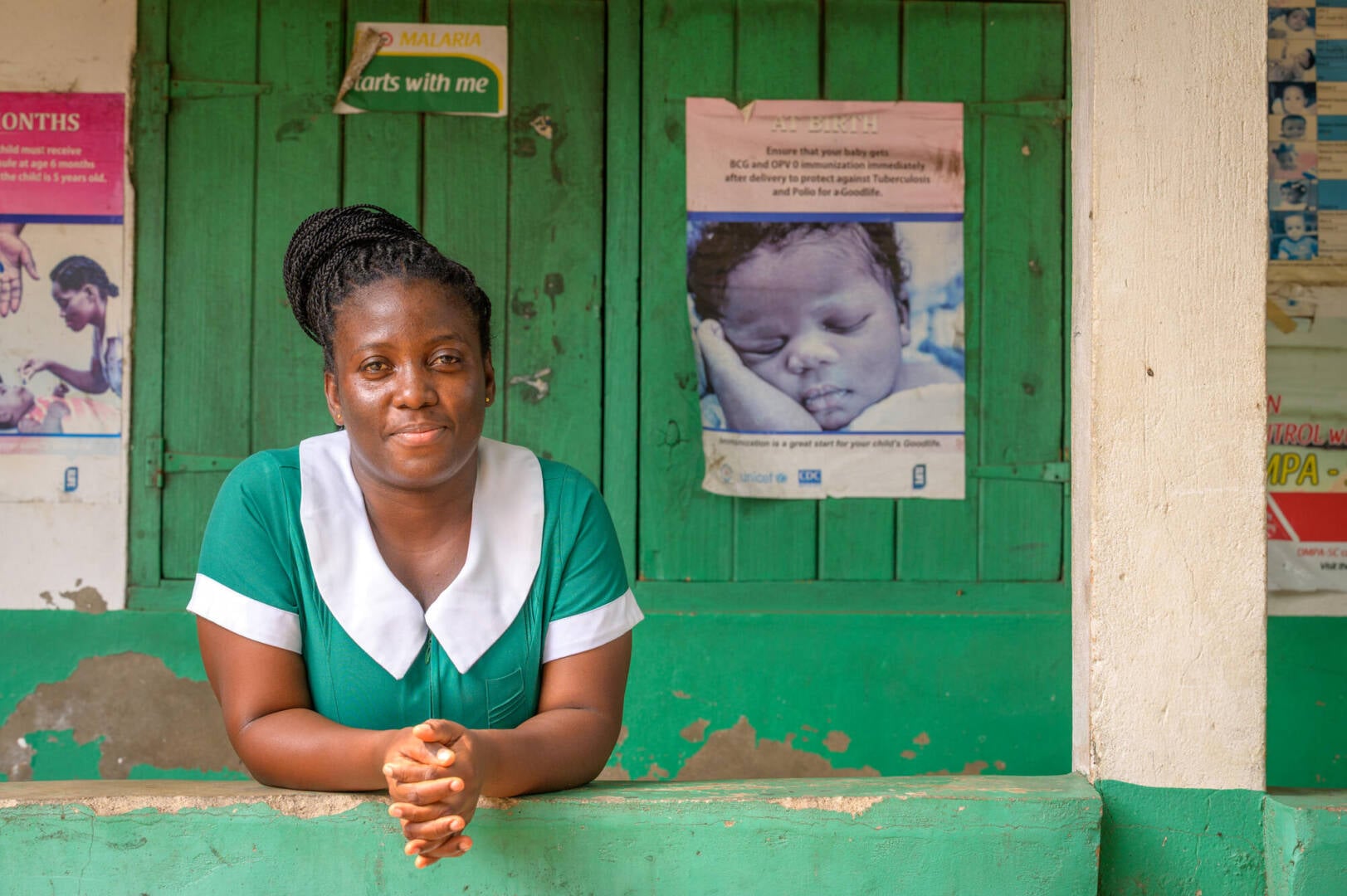 A woman leans on a short wall looking directly at the camera. On the wooden window covering behind her are posters of infants.
