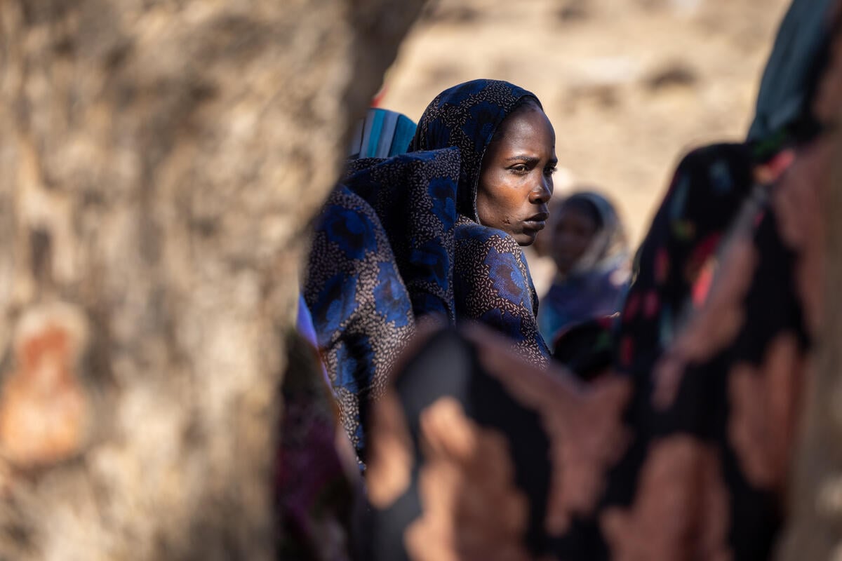 A woman looks to the side outdoors among other women and children. There is a tree trunk blurred in the foreground.