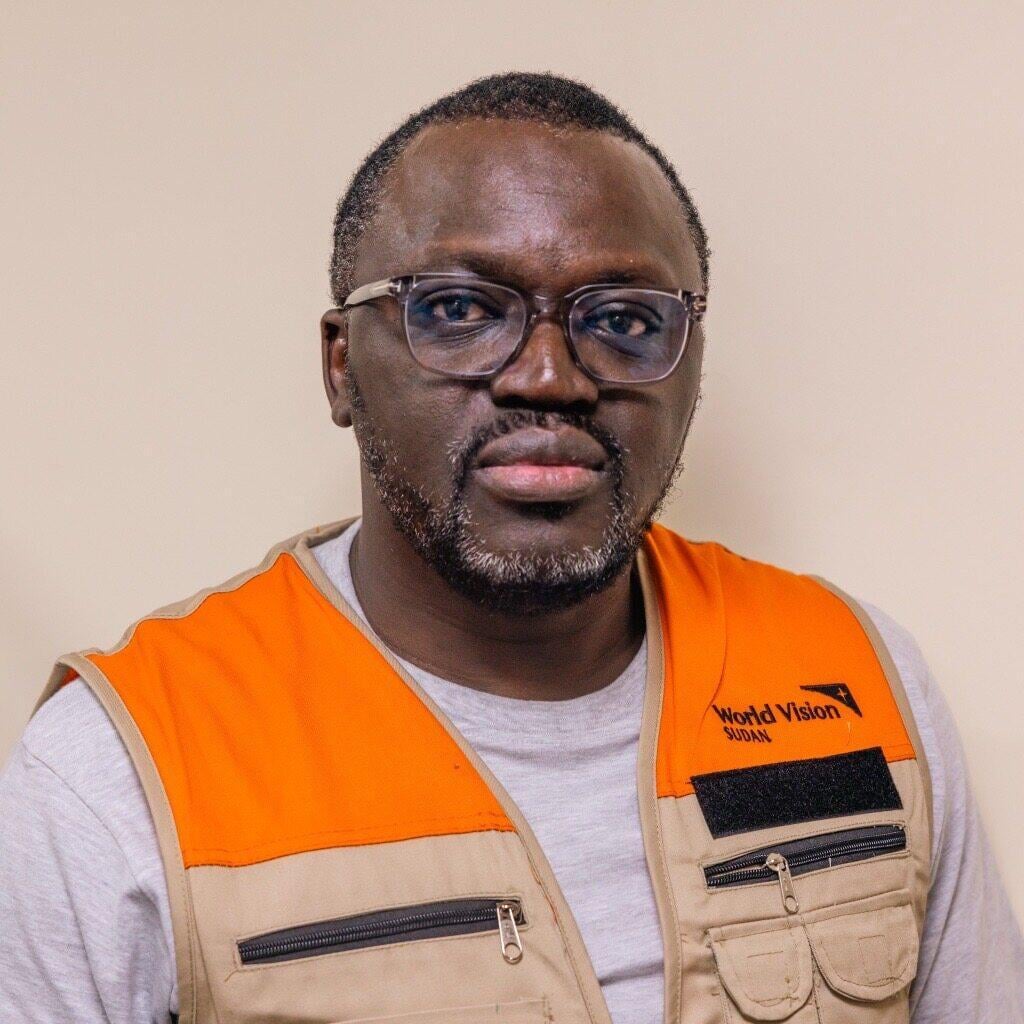 Portrait of a dark-complected man wearing glasses, a T-shirt, and a World Vision-branded vest.