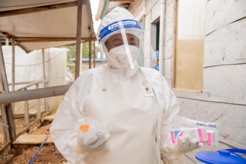 A health worker in protective gear and a face shield carries packages of medicine through an alley as another person in protective gear looks out from a doorway.