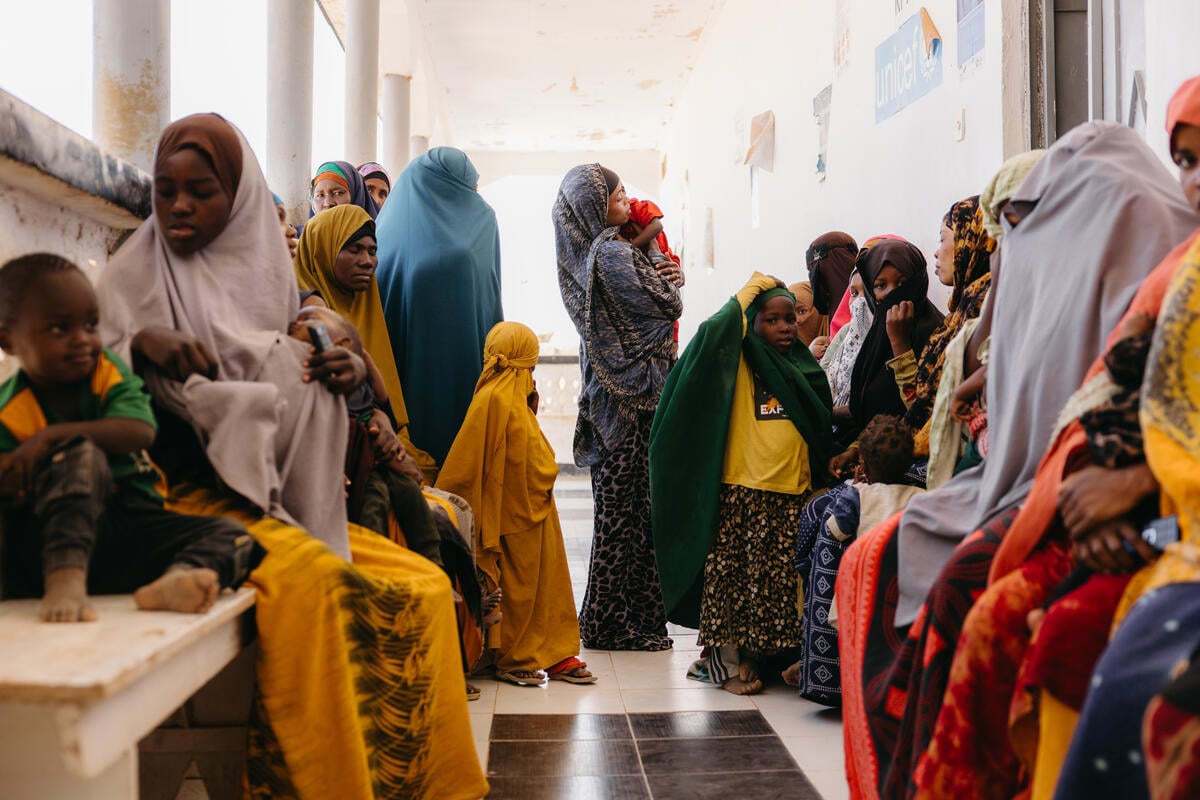 Women and girls in colorful dresses and headscarves wait in a clinic hallway.