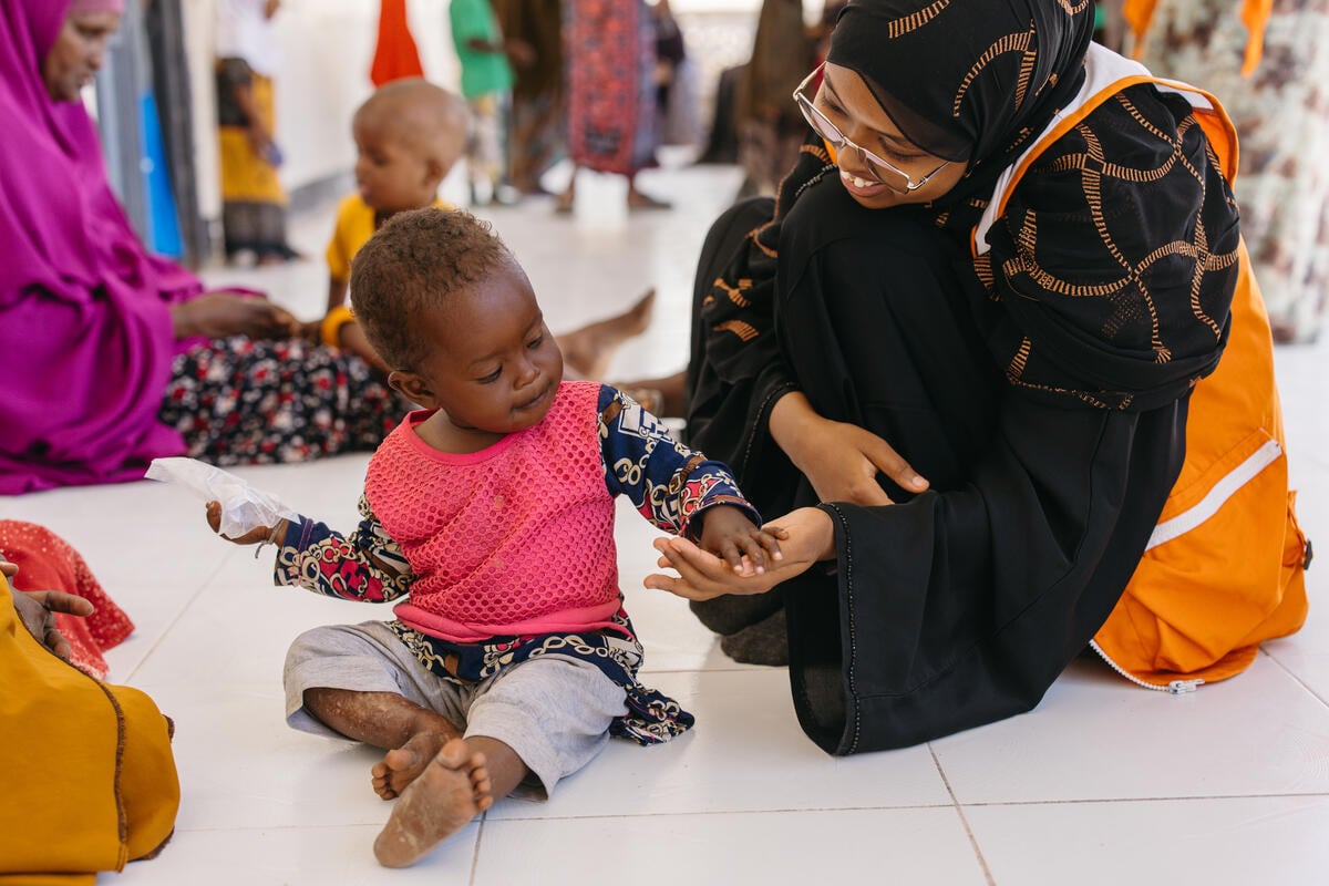 A woman wearing a headscarf looks at a young child seated beside her inside a health center, with other women and children in the background.