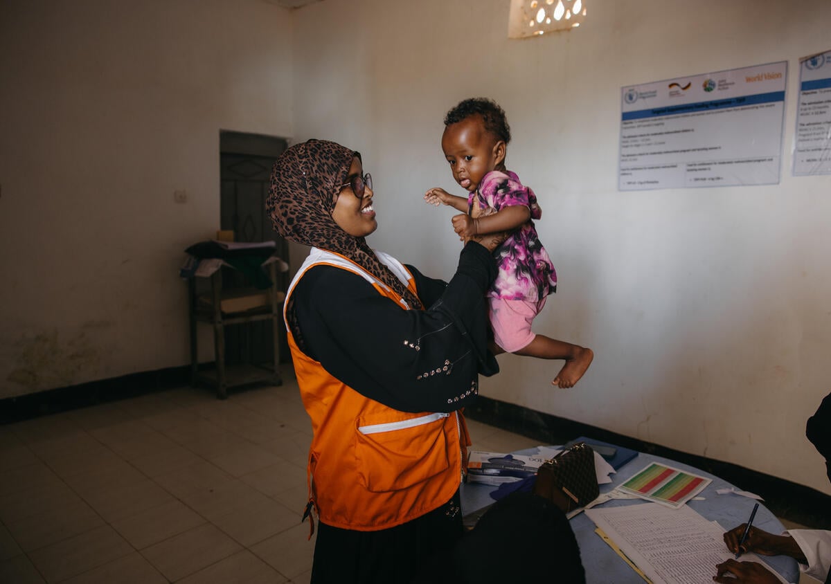 A health worker in a branded vest smiles as she raises a child with her arms.