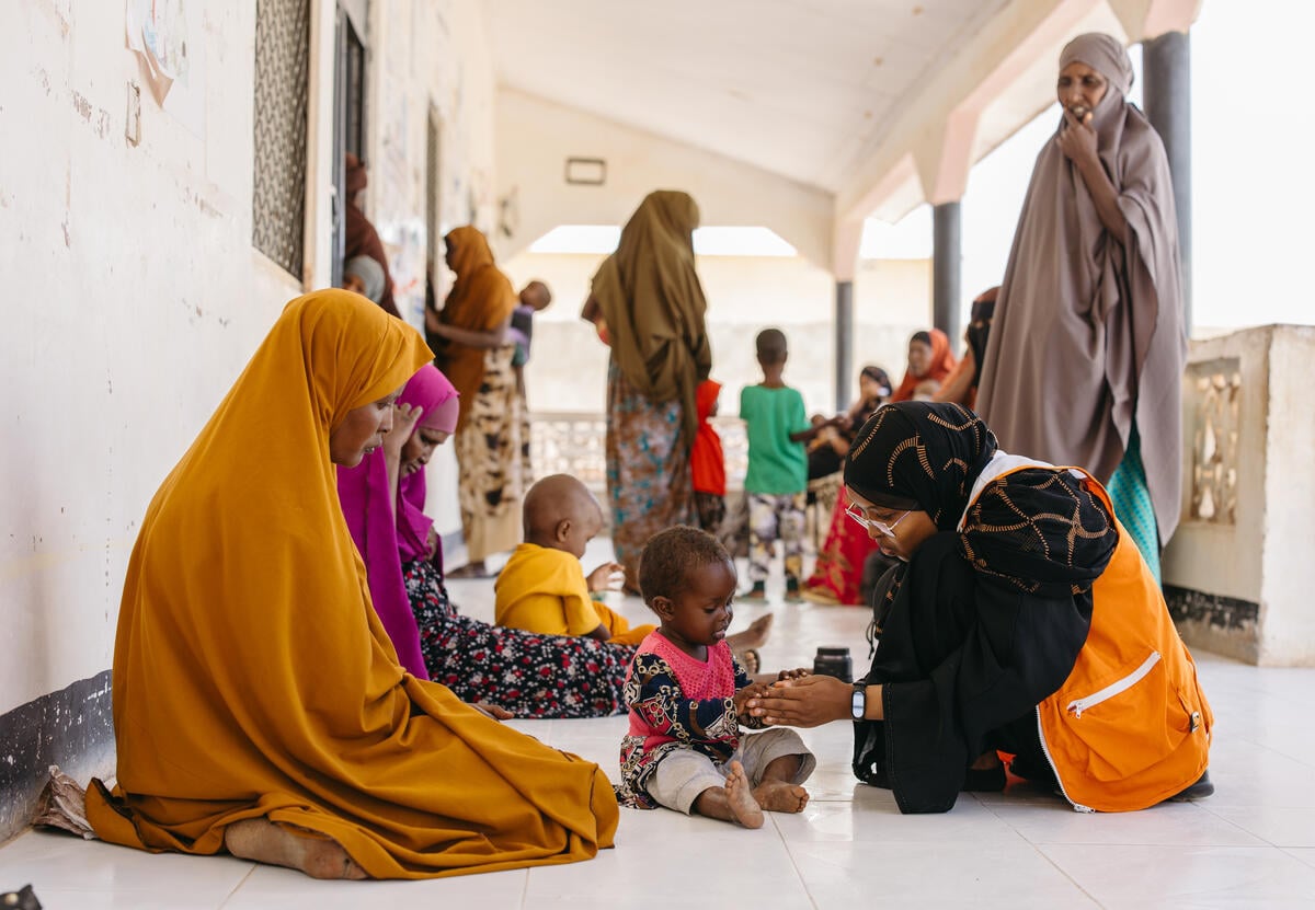Women dressed in colorful fabrics sit on the floor with children. One aid worker interacts with a young child.
