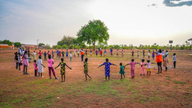 Dozens of children in colorful clothing stand in a wide circle, holding hands in an open field under a bright sky with a large tree in the distance.