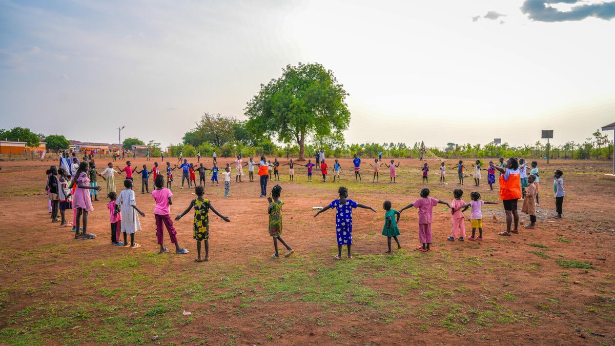 Dozens of children in colorful clothing stand in a wide circle, holding hands in an open field under a bright sky with a large tree in the distance.