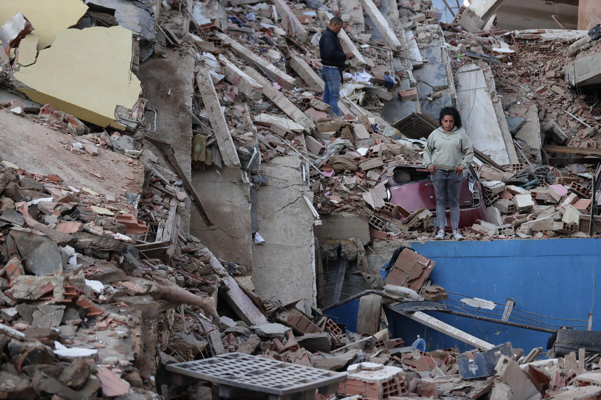 Two people stand amid piles of crushed concrete and debris that was once an apartment building.