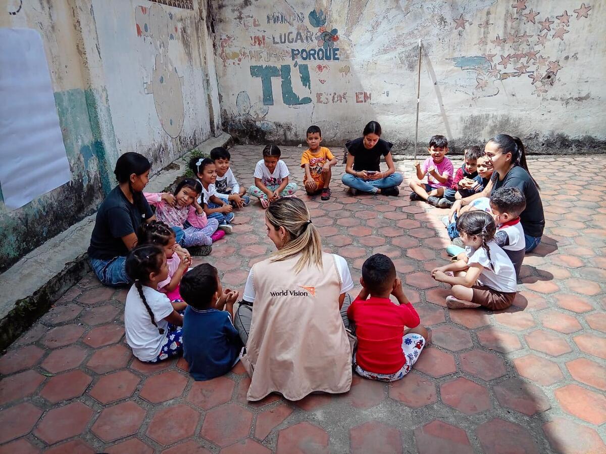 A World Vision aid worker sits with smiling children and two women in an open building, guiding a group activity in a safe place.