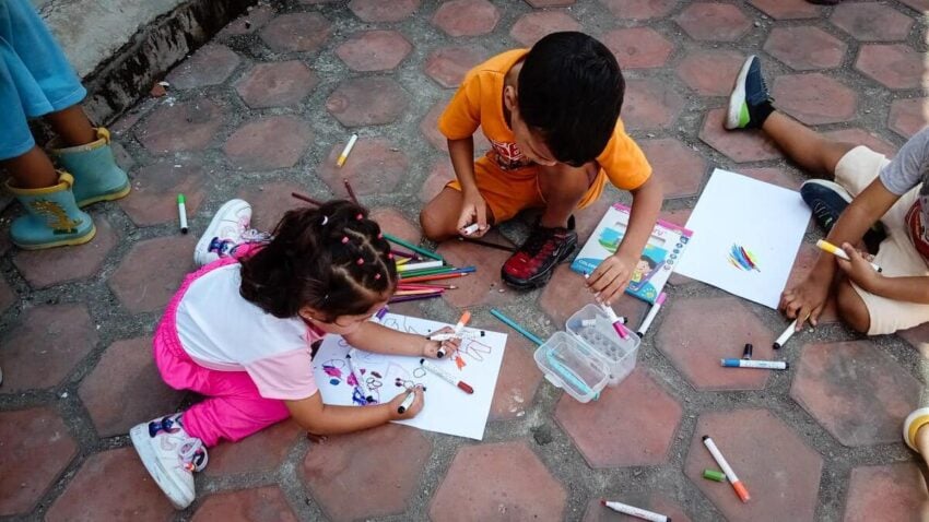 A young girl and boy sit on the floor inside a temporary shelter, coloring together during an activity. Markers and other children’s legs are visible in this serene scene.