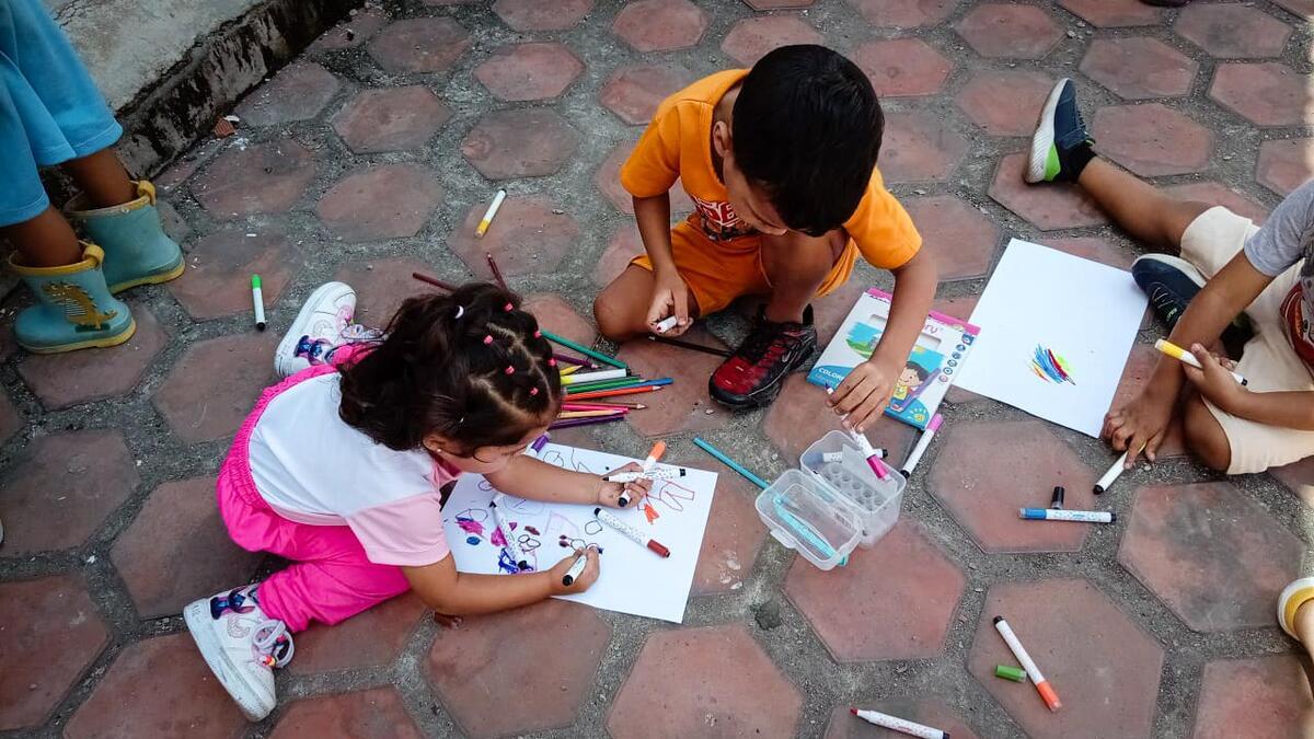 A young girl and boy sit on the floor inside a temporary shelter, coloring together during an activity. Markers and other children’s legs are visible in this serene scene.