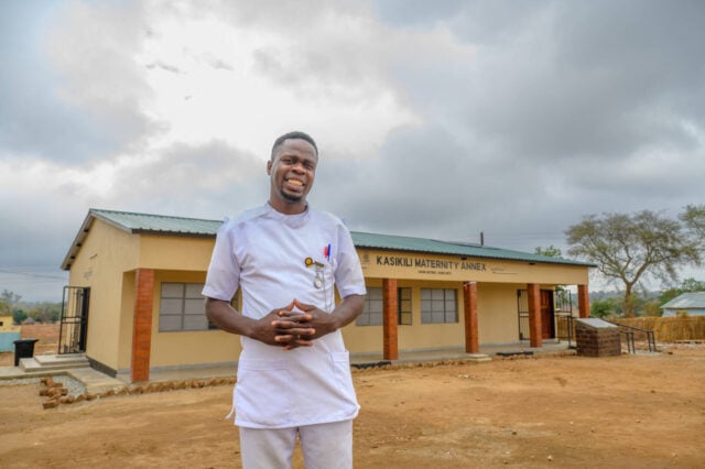 A man in a medical uniform stands in front of a building painted yellow with red brick columns.