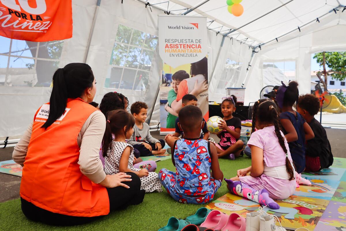 A young girl holds a white ball while sitting in a circle with other children and participating in an activity with them and an adult aid worker inside a colorful tent.