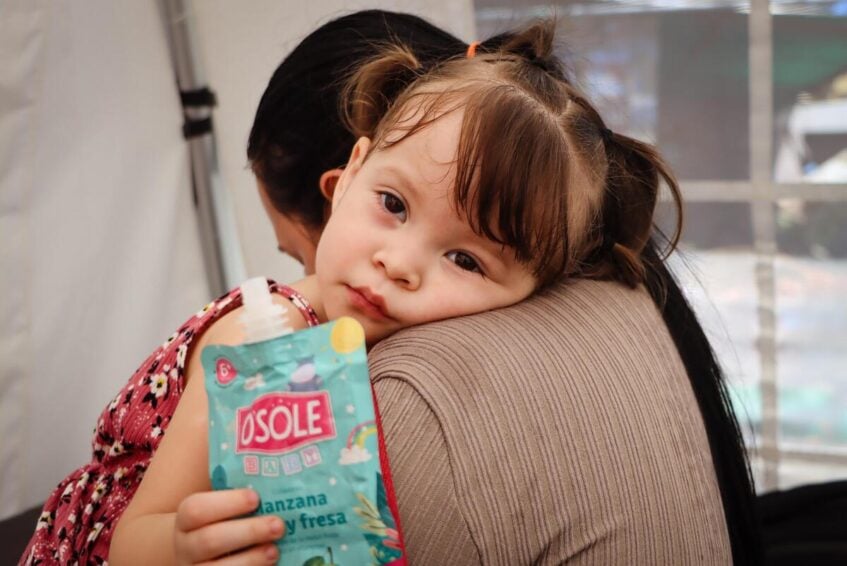 A young girl with pigtails holds a squeezable fruit pouch while resting on a woman’s shoulder and looks at the camera inside a temporary shelter.