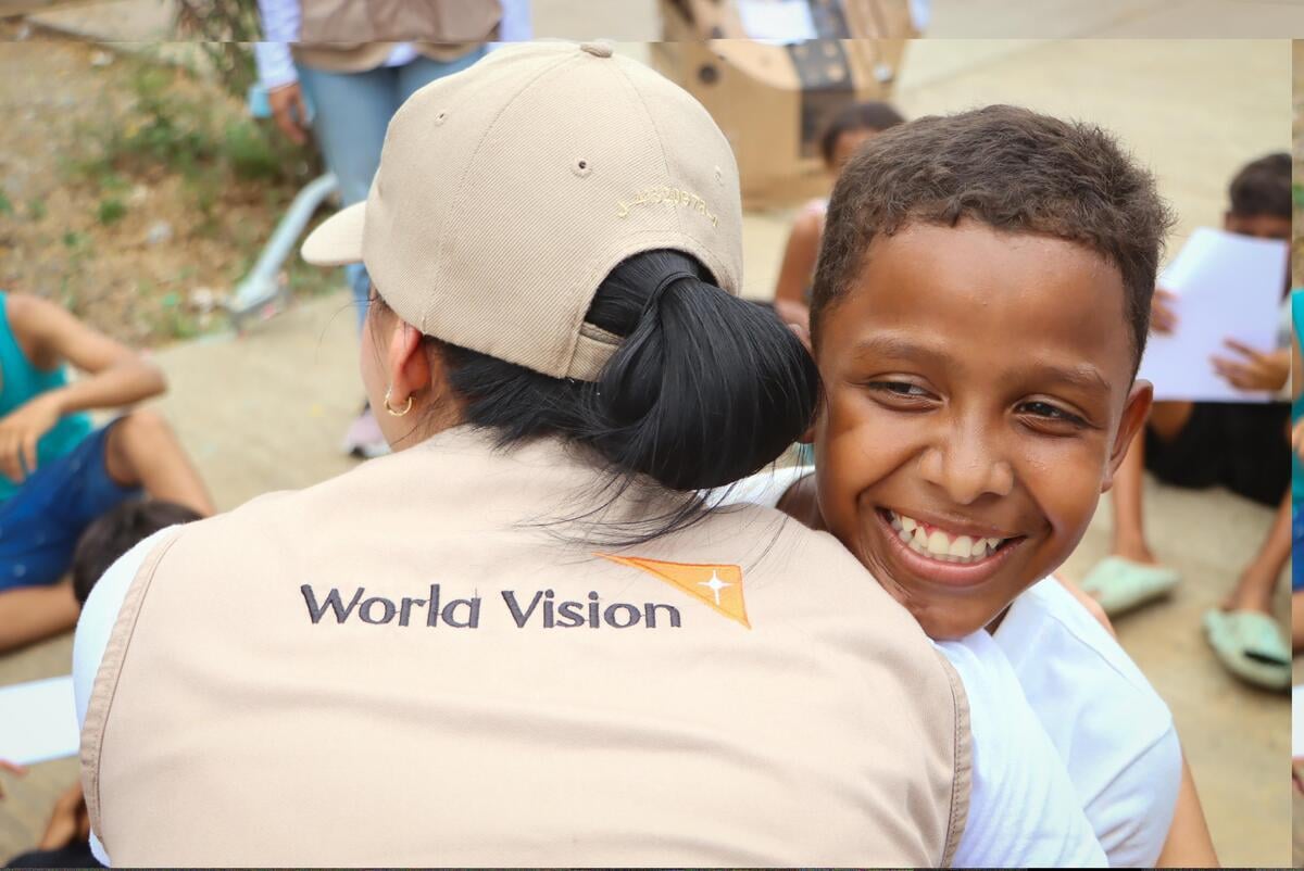 A child beams with a smile while hugging an aid worker in a World Vision vest outdoors, with&nbsp;other&nbsp;children blurred in the background.