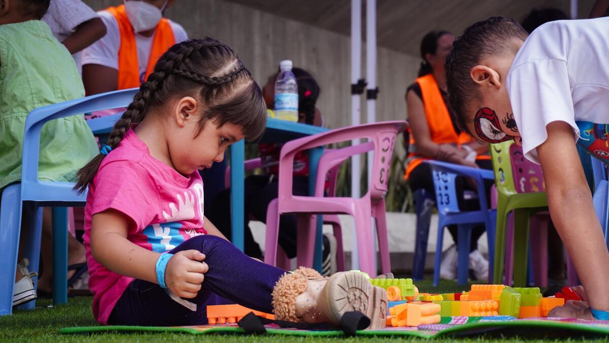 A young girl with braids sits on the floor across from a boy whose face is painted like Spider-Man. Colorful chairs are arranged behind them inside a shelter setting.