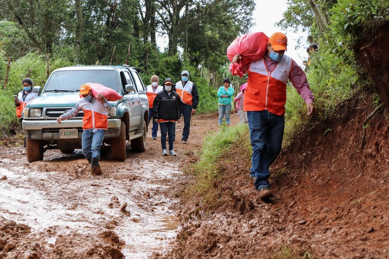 Men carry heavy bags while walking through mud on a rutted dirt road. There is a truck and people behind them as well as many trees.