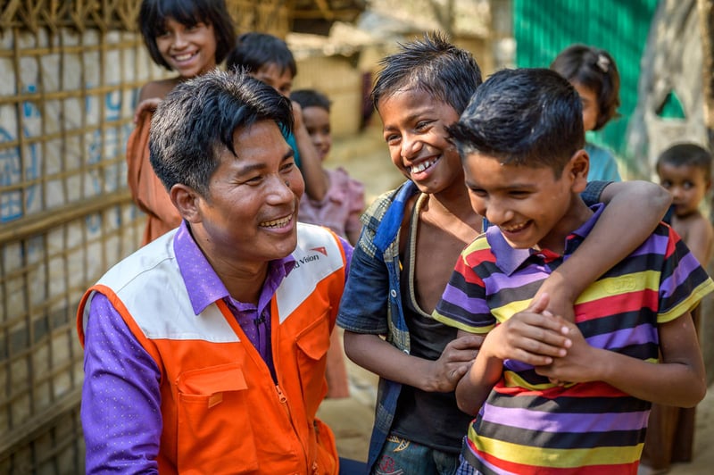 A man in an aid worker vest squats down to children&rsquo;s height level to laugh and talk with two boys, one of which has his arm around the other. Other children smile in the background.