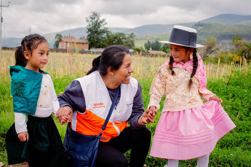 A woman in an aid worker vest kneels on the ground while holding the hands of two young girls wearing local dresses. There are mountains behind them.