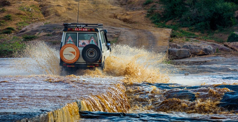 An SUV drives across a river, sending water splashing up about half the height of the vehicle. To the right of the vehicle are major boulders and a small rapids.