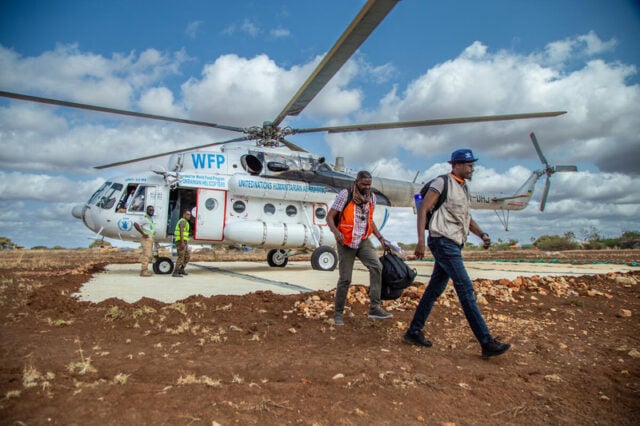 Two men walk away from a helicopter that is sitting on a cement pad surrounded by dirt as far as you can see. The sky is bright blue with large, puffy clouds.