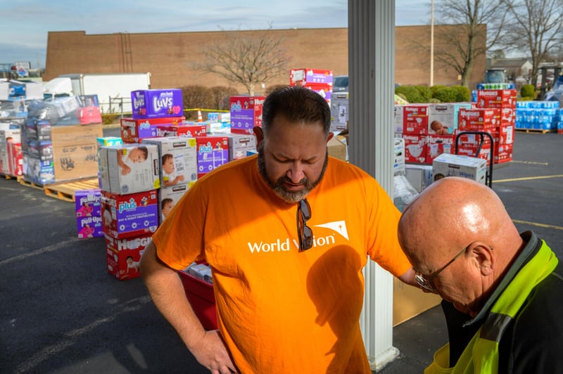 Two men have their heads bowed and eyes closed. One man has his hand on the other&rsquo;s shoulder, and there are boxes of diapers piled high behind them.