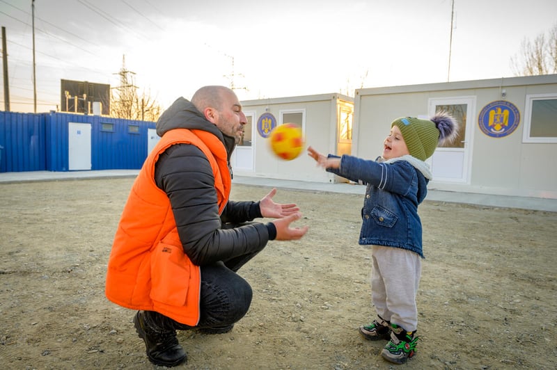 A man holds out his hands to catch a soccer ball that a child is throwing at him. The child is laughing, and both are dressed in warm winter clothes. There are buildings behind them.