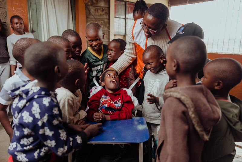 An aid worker stands above a child smiling and seated in a wheelchair. They are surrounded by other children.