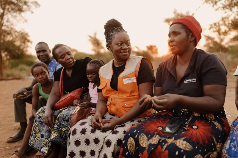 A woman in an aid worker vest looks at another woman as she speaks. Other women and children sit next to them also looking at the woman speaking. There are trees behind them.