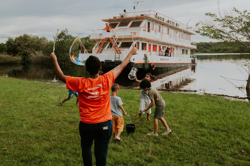 A woman holds up her hands as children play in front of her. A boat sits in the river behind them. There are trees on the other bank of the river.