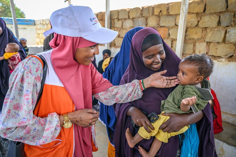 A woman in an aid worker vest over a dress touches the chin of a baby, who is being held by its mother. Both women look at the child lovingly. There are people and a brick wall behind them.