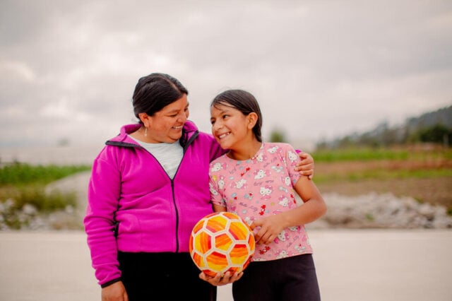 A woman smiles with her arm around the shoulder of teenage girl, who is holding a soccer ball and looking at the woman.