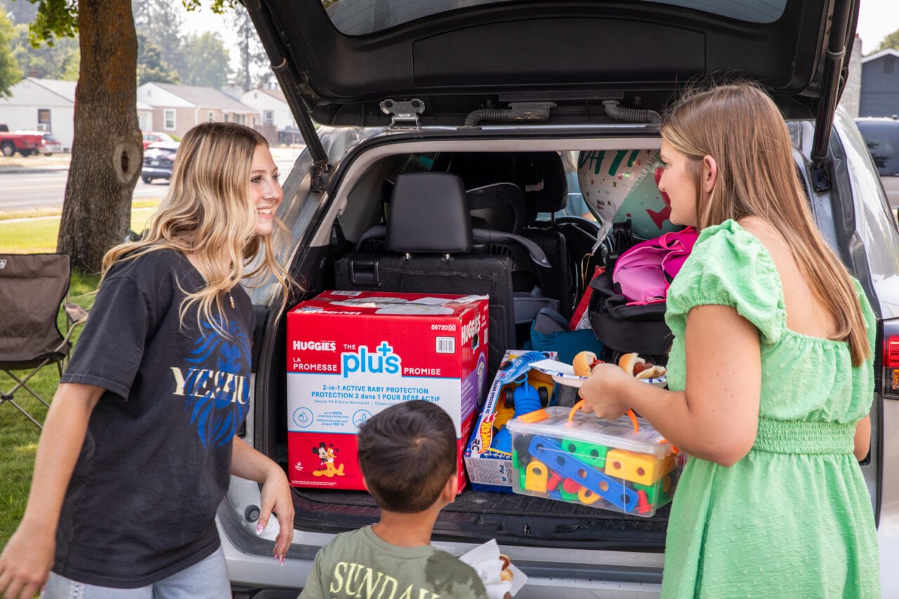 A young boy stands with two women as relief supplies are distributed. An open truck is stocked with a large box of diapers, toys, and other aid items.