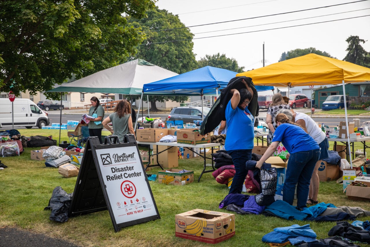 People gather at an aid distribution site with tables, tents, clothes, boxes of food, supplies, and a sign reading "Disaster Relief Center."