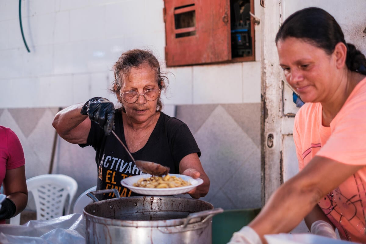 A woman wearing glasses and a T-shirt places food onto a plate from a large stock pot as another woman with her hair pulled back reaches for an item.