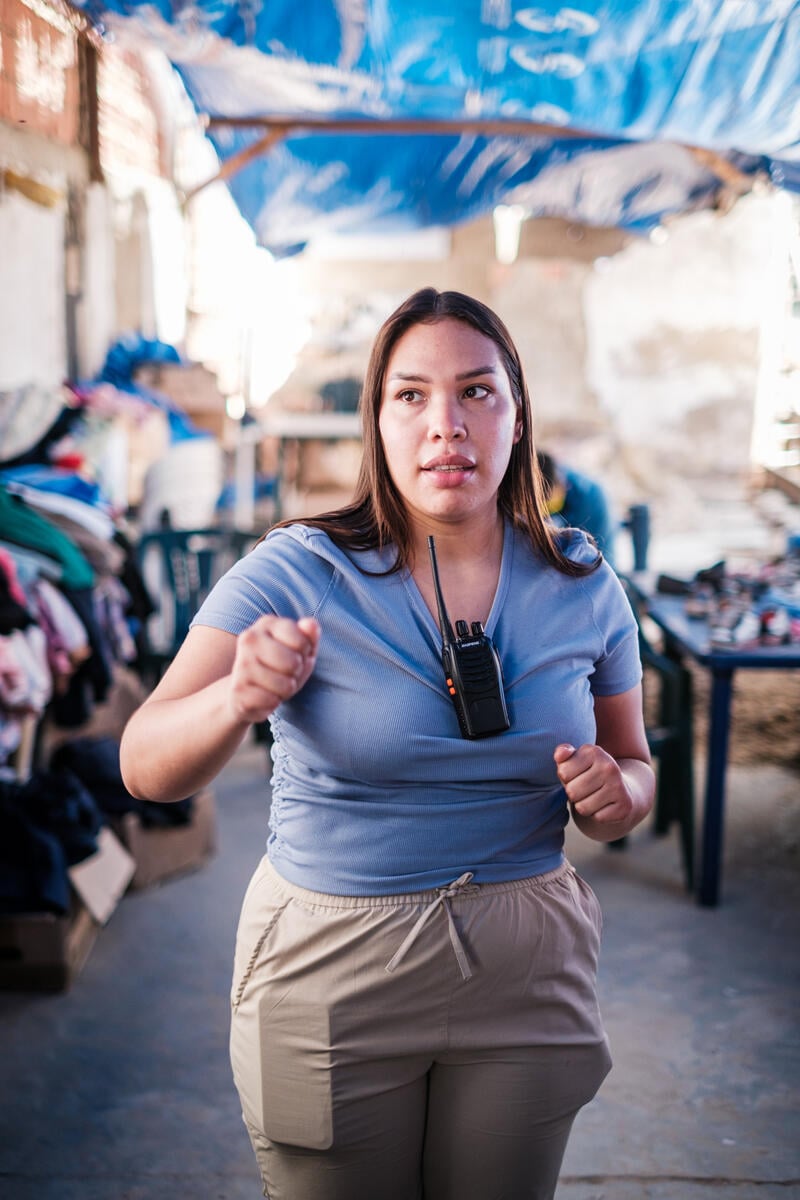 A woman with long hair glances to the side as she walks toward the camera. Tables filled with folded clothes are blurred in the background. Overhead, tarps frame the photo.