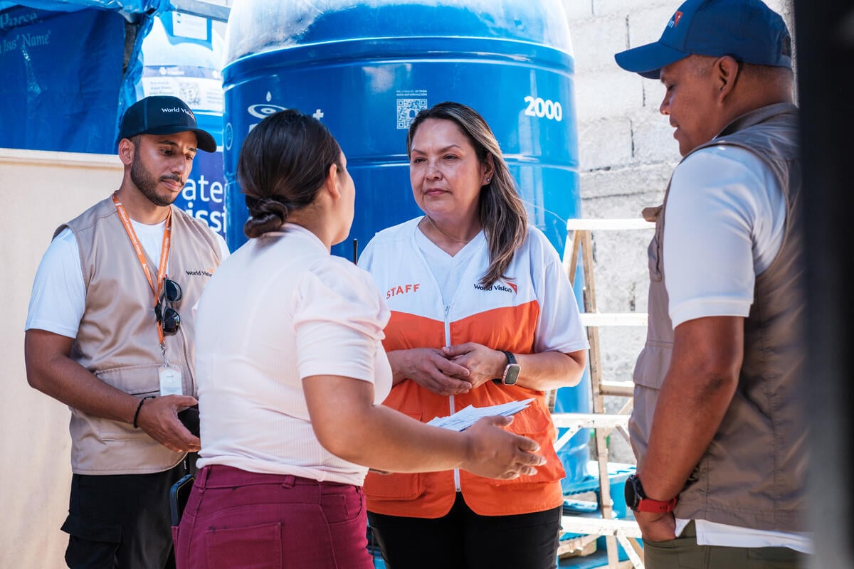 Three World Vision aid workers and a woman stand in a circle in conversation. A ladder and large water container are visible in the background as daylight streams from above.