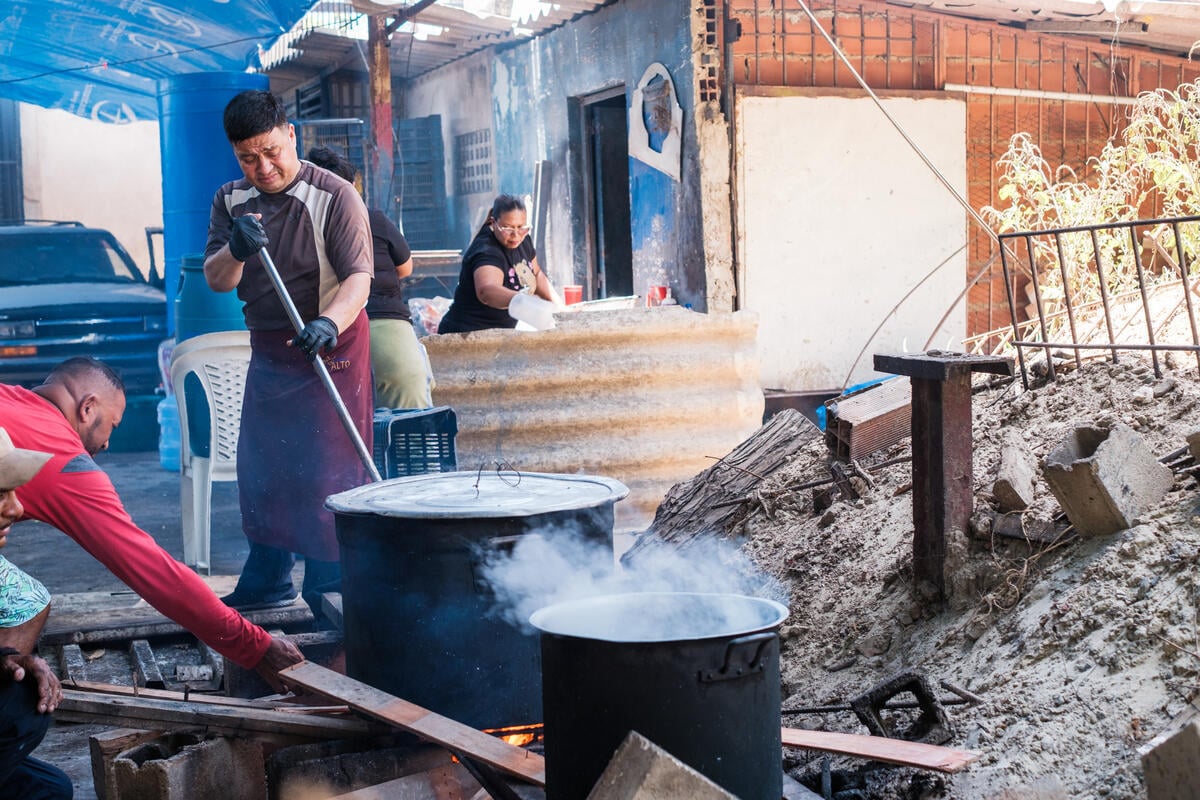 One man stands with a metal rod stoking an open fire, while another man adjusts a plank nearby. Two large pots sit over the flames. Rubble lines the side of the damaged structure. A woman is in the background.