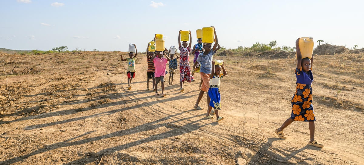 Girls and boys carry water containers on their heads through a dry, parched landscape under the bright sun.
