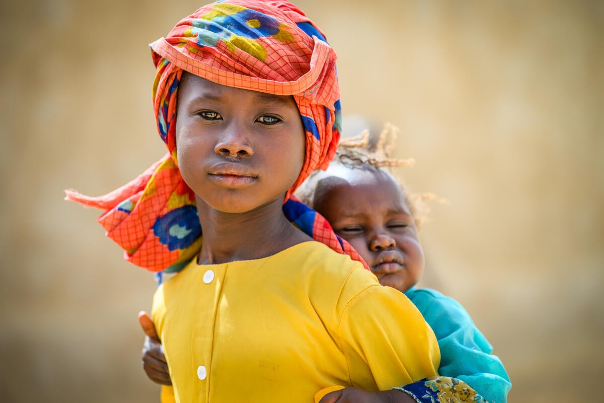 A young girl with a steady gaze carries a baby on her back.