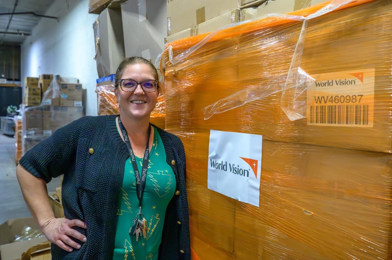 A woman leans against a pallet of essential supplies sent by World Vision. Her hand is on her hip and she smiles for the camera.