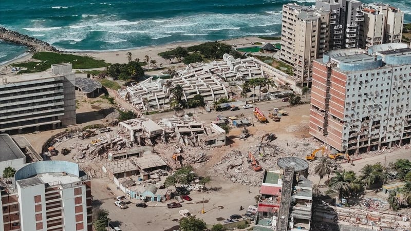 An aerial view of collapsed buildings and severely damaged infrastructure. The buildings run along a beach with blue water.