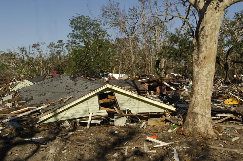 A roof of a house sits on the ground after everything below it had collapsed. There is rubble all around and a tree in the foreground.