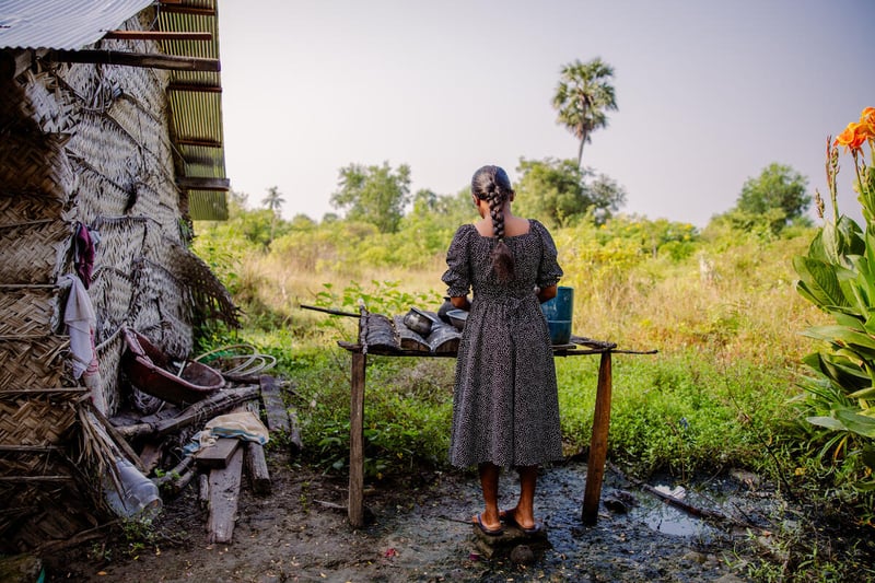 A woman stands with her back to the camera as she washes dishes at a table made from sticks and tin. The house next to her is cobbled together with grasses, ties, and plastic with a thin metal roof.