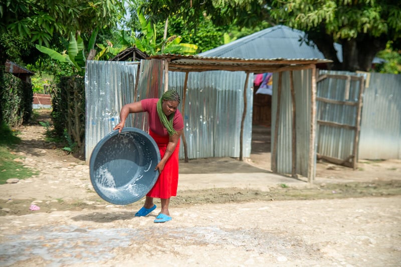 A woman empties a large basin of soapy water on the ground outside a home made of sticks and tin.