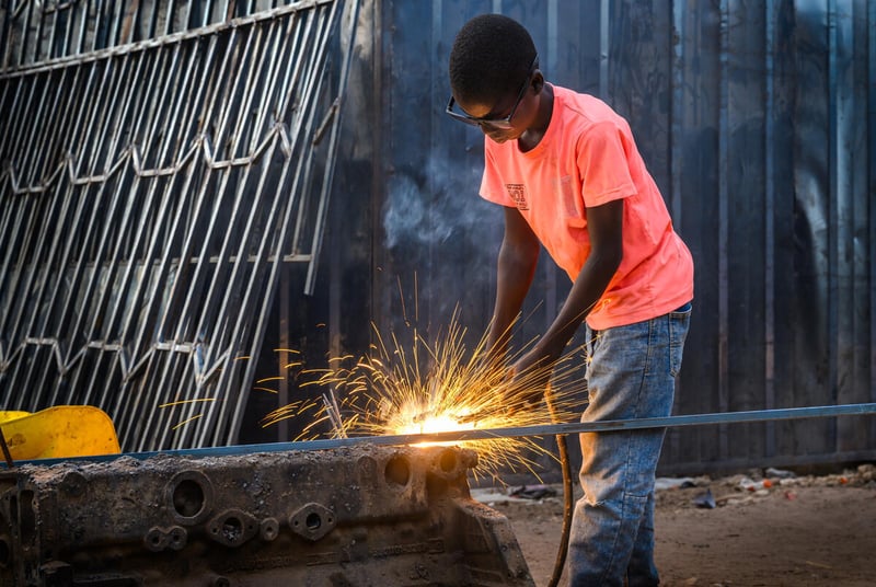 A boy leans over a piece of a metal while using a tool that causes sparks to fly.