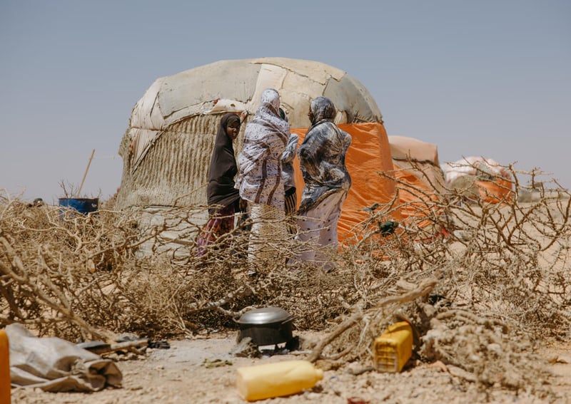 Women stand outside a makeshift hut talking. The ground is dry and there are dry branches in the foreground.